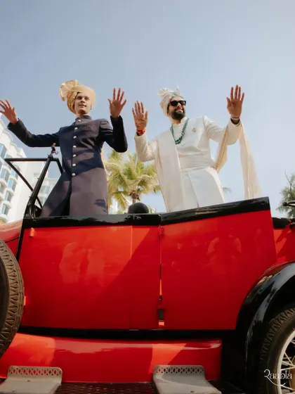 The groom and his baraat making a grand entrance in a vintage red car, waving to the crowds.