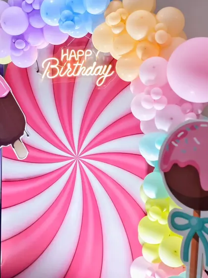 A fun and colorful candy-themed backdrop with a swirling lollipop design, an ice cream bar cutout, and a 'Happy Birthday' neon sign.