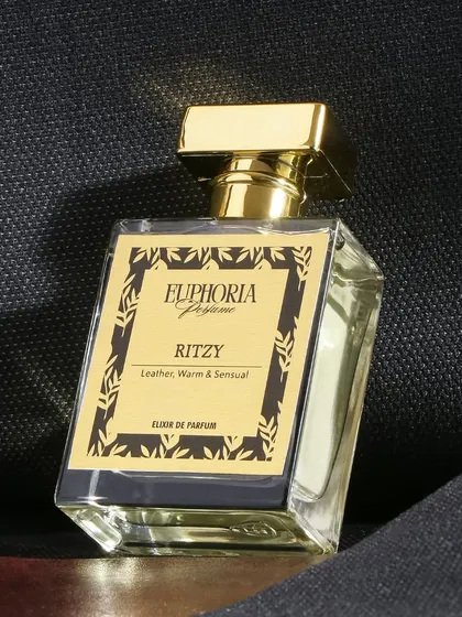 A sleek and minimalist shot for 'Ritzy' perfume against a textured black background. The focus is entirely on the luxurious gold bottle, conveying a sense of modern, understated elegance.