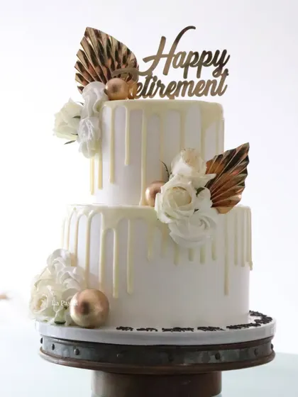 Celebrating a milestone with this two-tier retirement cake. The design is clean and sophisticated, with a white chocolate drip, white roses, and metallic gold palm spears for a modern, celebratory look.