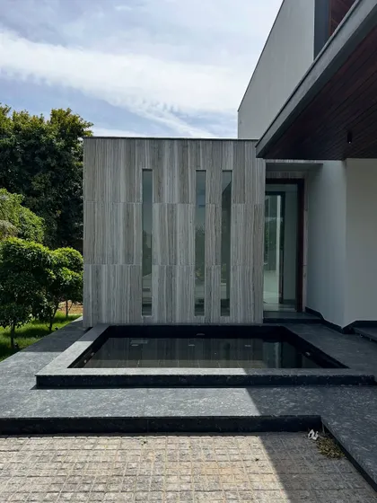 This close-up shows the serene water body at the entrance of Rosewood Farmhouse. The feature is bordered by dark stone and sits beside a wall of vertically laid grey stone with slit windows, creating a beautiful interplay of textures and reflections.