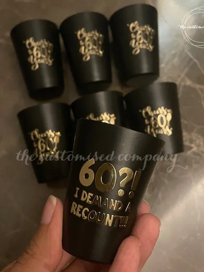 Cheers to 60 years! These custom black shot glasses are perfect for a milestone birthday party, featuring cheeky phrases like "60?! I DEMAND A RECOUNT!" in elegant gold lettering.