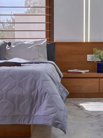 A calm and restful bedroom corner. The low wooden bed frame extends into a convenient bedside bench, and the large windows with slatted blinds allow for soft, diffused sunlight.