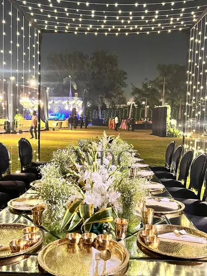 An elegant long-table dining setup, illuminated by a canopy of fairy lights. The table is adorned with a runner of white flowers and set with golden 'thali' plates for a touch of tradition.