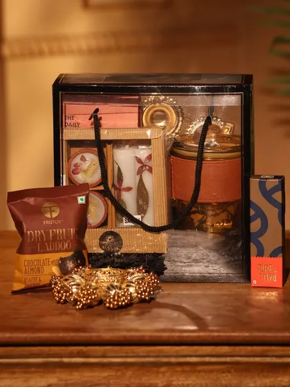 This elegant Diwali gift set is presented in a transparent bag with black accents. It includes dry fruit laddoos, a decorative candle, and other festive treats.