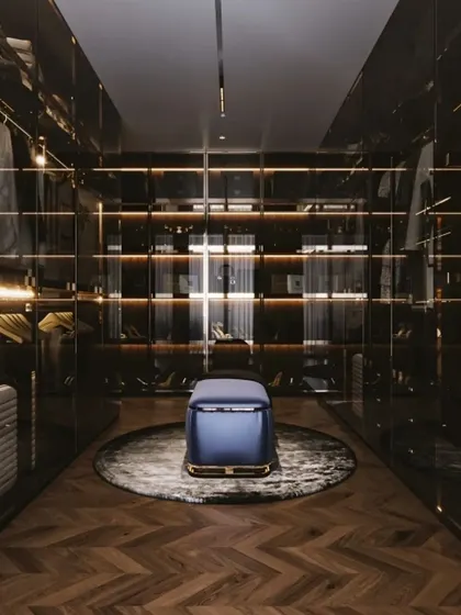 An ultra-luxurious walk-in closet with dark wood, smoked glass doors, and warm accent lighting. The central ottoman and herringbone flooring complete the sophisticated and exclusive feel.