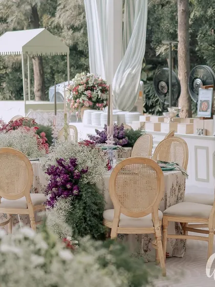 This outdoor dining setup features classic rattan chairs and custom floral tablecloths. The tables are surrounded by lush arrangements of purple statice and baby's breath, creating a beautiful garden party feel.