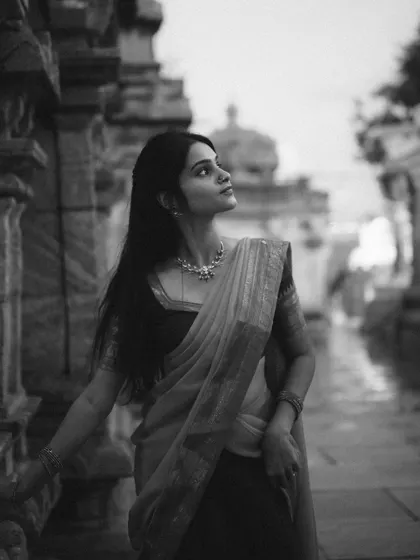 A black and white portrait of a model in a saree, taken at a temple. The monochrome treatment emphasizes the textures, shadows, and timeless elegance of the scene.