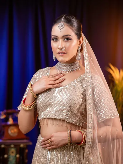 A graceful and polished look featuring a model in a shimmering silver-white lehenga. The soft studio lighting highlights the subtle glow of the makeup and the intricate details of the diamond necklace and traditional bangles, creating a perfect sangeet or reception look.