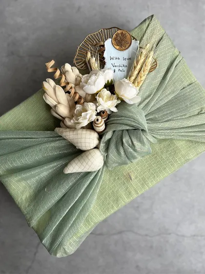 An exquisitely wrapped gift using a sage green fabric in the Furoshiki style. The knot is adorned with a bouquet of shola wood flowers, dried wheat, and other natural elements, all tied together with a handwritten tag.