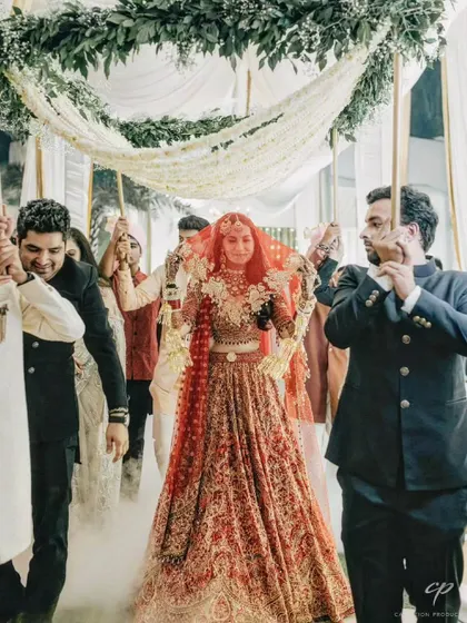 The bride makes her grand entrance under a phoolon ki chadar made of lush greenery and white flowers, held by her brothers, creating a classic and emotional bridal procession moment.