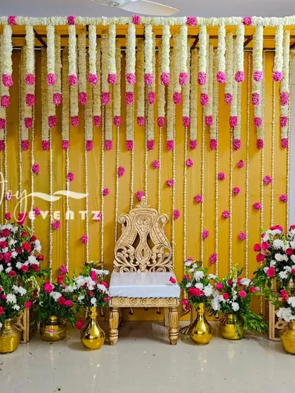 We designed this elegant stage with hanging garlands of tuberose and pink carnations against a bright yellow backdrop. The setup includes a golden chair and matching brass flower vases for a traditional ceremony.