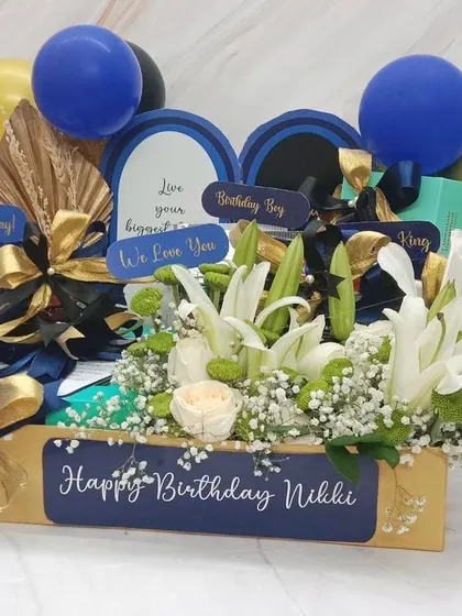 A stylish and modern birthday hamper for 'Nikki'. The blue and gold theme is carried through from the balloons and ribbons to the personalized crate, filled with white lilies and roses.