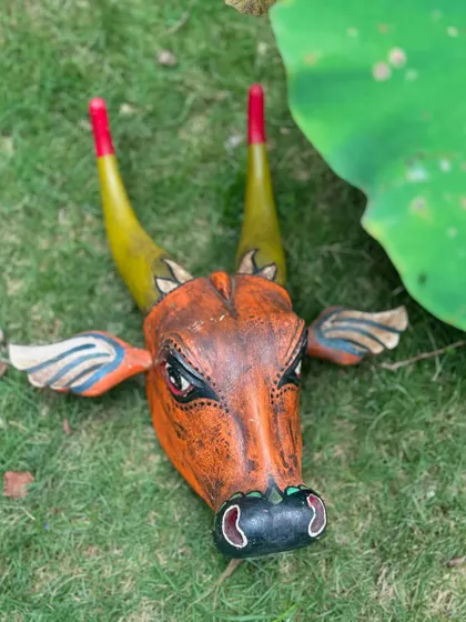 Another view of the hand-painted wooden bull head, resting on the grass. The bright, festive colors are inspired by the grand cattle festivals of Kerala, bringing a piece of that tradition to your home.