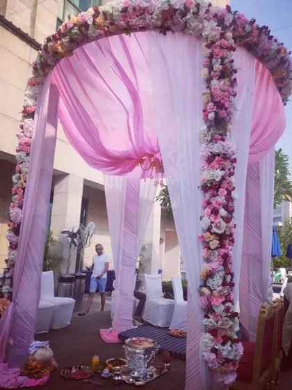 A lovely poolside mandap setup. The circular structure is draped in pink and white fabric and adorned with cascading pink floral arrangements, creating a romantic setting for the pheras.
