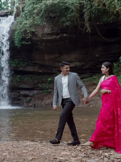 A couple holds hands and walks along the edge of a stream with a waterfall behind them. This photo captures a playful and romantic moment in a beautiful natural setting.