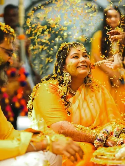 A candid moment of pure joy during a Haldi ceremony. The woman's face is covered in turmeric paste as she is showered with flower petals, her laughter lighting up the frame.