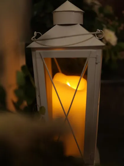 A close-up of a white lantern holding a pillar candle. This classic decor item adds a touch of rustic charm and warmth to any setting.