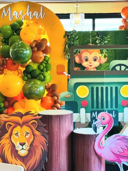 A detailed shot of the jungle party decor, focusing on the lion prop and the personalized jeep. The image highlights the rich textures, from the wood-patterned plinths to the green balloon garland, creating an authentic safari feel.