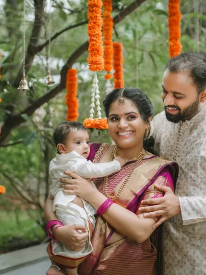 A lovely family portrait taken outdoors during a naming ceremony. The parents embrace their child under hanging marigold flower arrangements, creating a warm and traditional photograph.