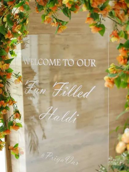 A lovely acrylic welcome sign for a "Fun Filled Haldi," framed by delicate orange blossoms.