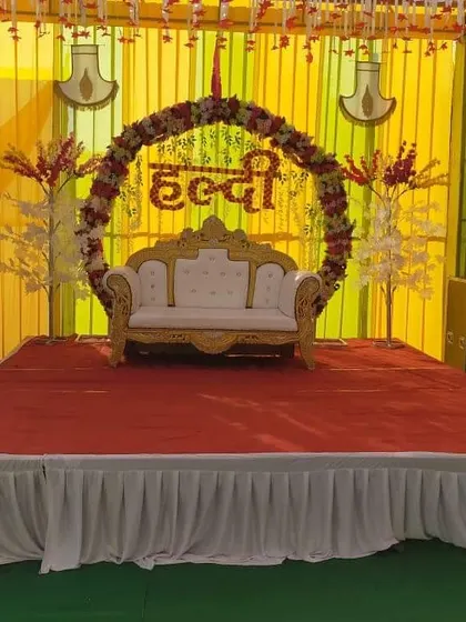 A traditional Haldi stage setup with a yellow and white color scheme. The stage includes a floral ring with the word 'Haldi', a royal sofa, and a sound system.