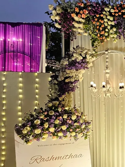 A close-up of a floral arrangement on a welcome stand. The combination of purple and white flowers with soft fairy lights creates a dreamy and inviting detail.