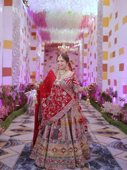 A full-length portrait of a radiant bride in her magnificent red and gold lehenga, standing in a beautifully decorated wedding hall.