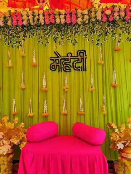 A Mehendi backdrop with a lime green curtain, hanging floral buds, and a 'Mehendi' sign. The decor is simple, focusing on a fresh color palette.