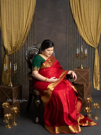 A beautiful portrait of a mother-to-be seated in a regal chair. She is wearing a rich red silk saree, looking down thoughtfully in a traditionally styled studio.