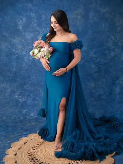A gentle and classic maternity pose. Holding a bouquet of flowers, the mother-to-be looks down at her bump with love, her profile highlighted against the rich blue backdrop.