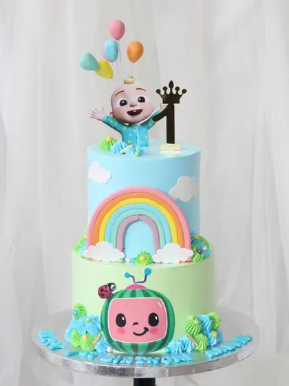 A bright and cheerful Cocomelon cake for a first birthday. This two-tiered design features the iconic Cocomelon character, a colorful rainbow, and the baby JJ character on top.