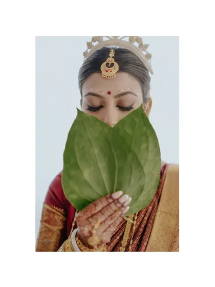 A playful yet traditional pose. The bride peeks over betel leaves, her eyes telling a story of anticipation and joy, a classic moment in Bengali wedding photography.
