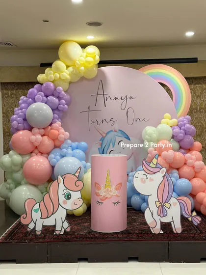 A dreamy pastel unicorn theme for a first birthday party. This setup includes a circular backdrop, cute unicorn cutouts, a rainbow accent, and a soft, cloud-like balloon garland.
