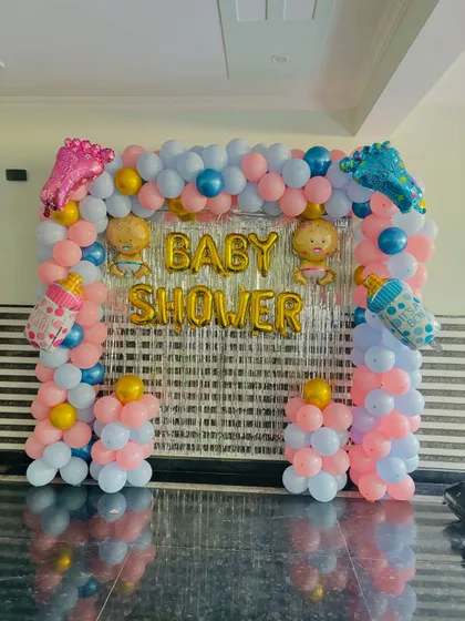 A classic baby shower backdrop with a balloon arch in pink and blue, a silver tinsel curtain, and baby-themed foil balloons.