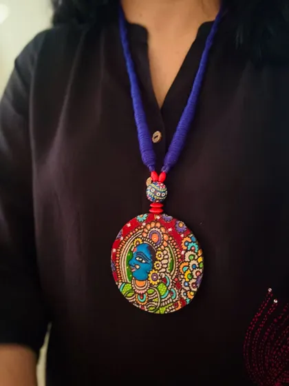 A beautiful hand-painted face in the Kerala mural style, with vibrant colors and expressive eyes. This piece is strung on a simple blue cord to let the artwork shine.