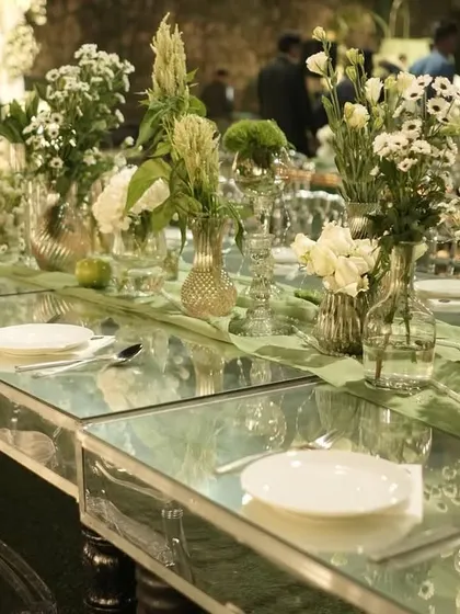 A close-up of the mirrored dining table, reflecting the floral centerpieces and the green drapes above.