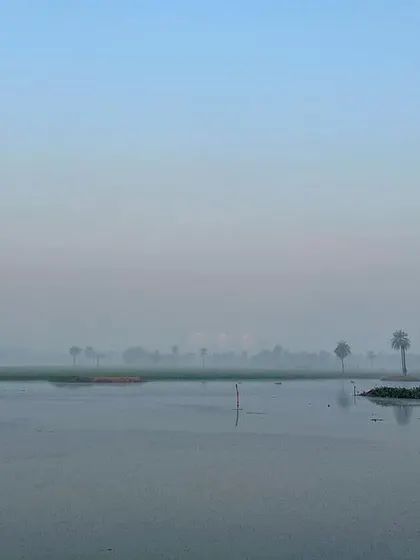 A wide, minimalist landscape of the Surajpur wetlands on a foggy morning. The calm water and distant treeline create a feeling of immense peace and space.