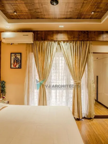 The bedroom is filled with natural light, enhanced by light-colored curtains and white wardrobes. A wooden false ceiling and matching floor add warmth and continuity to the design.