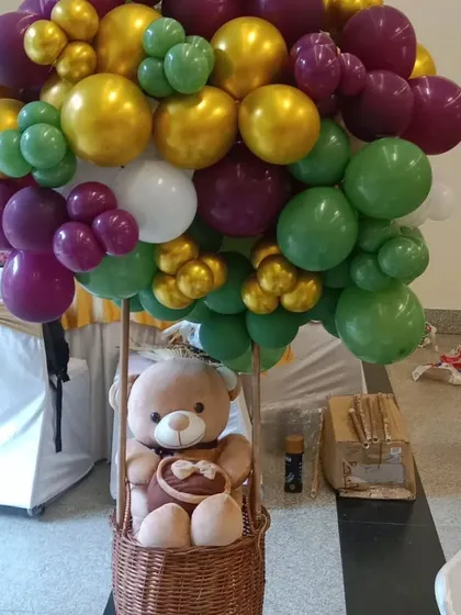 A teddy bear in a hot-air balloon centerpiece. I use a wicker basket and a large bunch of balloons in green, purple, and gold to create this popular and whimsical decorative piece.