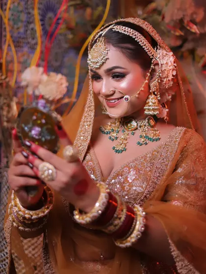 A candid moment of a bride admiring herself in a hand mirror. This close-up shot captures her joyful expression and the stunning details of her golden lehenga and green jewellery.
