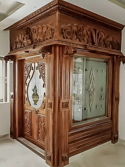 A magnificent, fully enclosed pooja room made of carved wood and etched glass. The elephant carvings on the top panel are a symbol of good luck and prosperity.