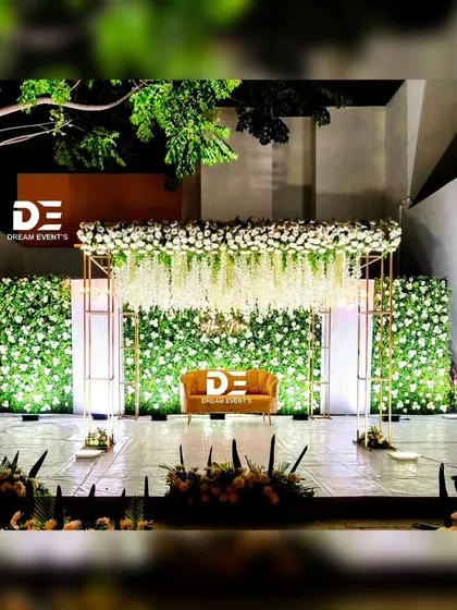 A modern and chic outdoor reception stage design. I used a full green floral wall as the backdrop, accented with hanging white wisteria and a minimalist gold frame to create a fresh, garden-like ambiance.
