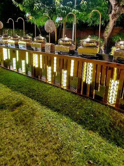 A beautifully lit rustic wooden buffet counter set up outdoors. The warm internal lighting and traditional brass serving dishes create a cozy and inviting atmosphere for an evening gathering.
