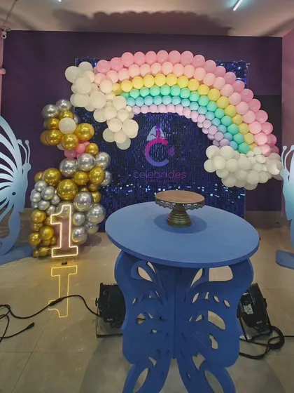 A rainbow and butterfly-themed first birthday party. The pastel balloon rainbow against a blue sequin wall and the butterfly-shaped tables are perfect for a little one's celebration.