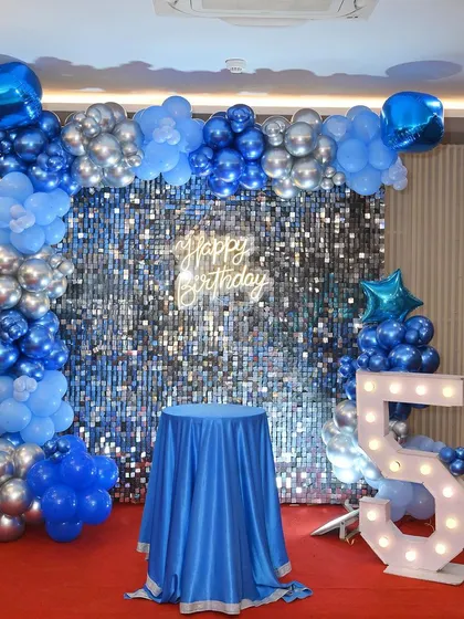 A simple yet elegant birthday backdrop with a blue and silver theme. The decor includes a silver shimmer wall, a blue balloon arch, and a marquee number '5'.