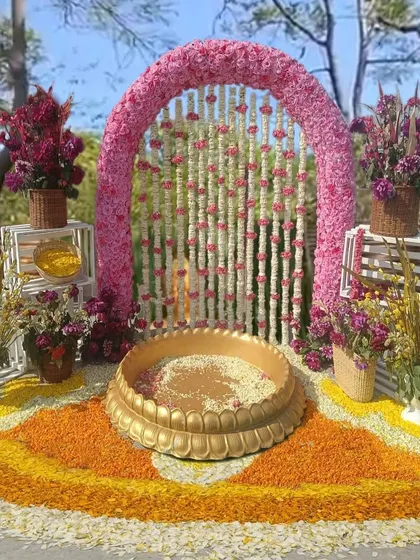 A beautiful Haldi ceremony setup with a pink floral arch and a traditional brass urli. The floor is decorated with a colorful rangoli made from fresh flower petals, adding to the festive look.