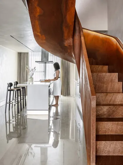 This view showcases the relationship between the sculptural staircase and the open-plan kitchen. The raw beauty of the Corten steel and wooden steps creates a powerful visual anchor in the home's otherwise understated and monochromatic design.