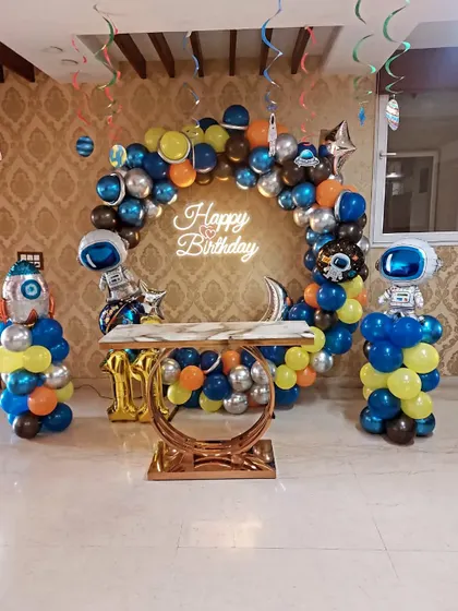 An out-of-this-world space-themed birthday party. The setup includes a balloon arch with metallic silver, blue, and orange balloons, astronaut foil balloons, and a matching cake table.