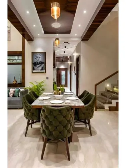 A view of a spacious dining area with tufted green velvet chairs and a marble-top table. The high ceiling with wood paneling and modern pendant lights adds to the room's grandeur.